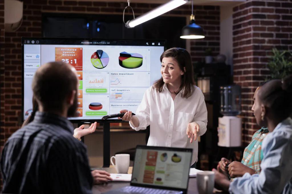 Smiling caucasian woman in startup office doing business presentation on big tv screen with charts